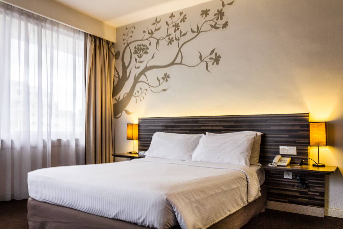 A hotel room with a large bed, two bedside tables with lamps, a wall mural, and a window with curtains.