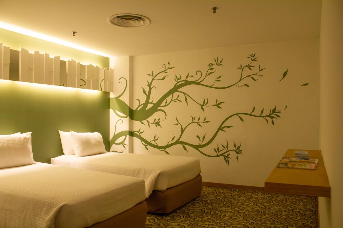 A hotel room with two beds, a green accent wall, a wall mural of a tree, and a patterned carpet.