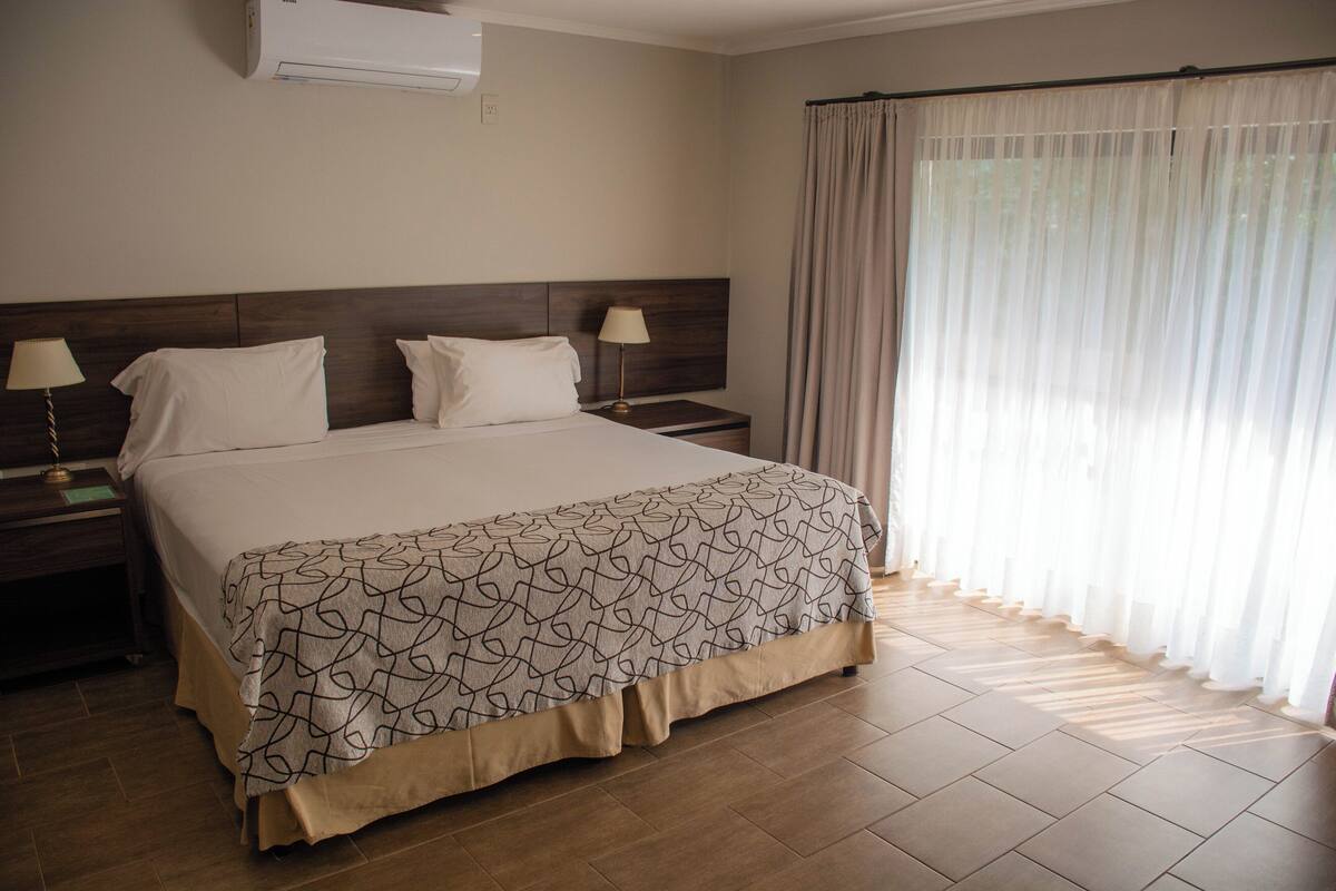 Habitación de hotel con una cama grande, dos mesitas de noche con lámparas, un cabecero de madera y una ventana con cortinas translúcidas.