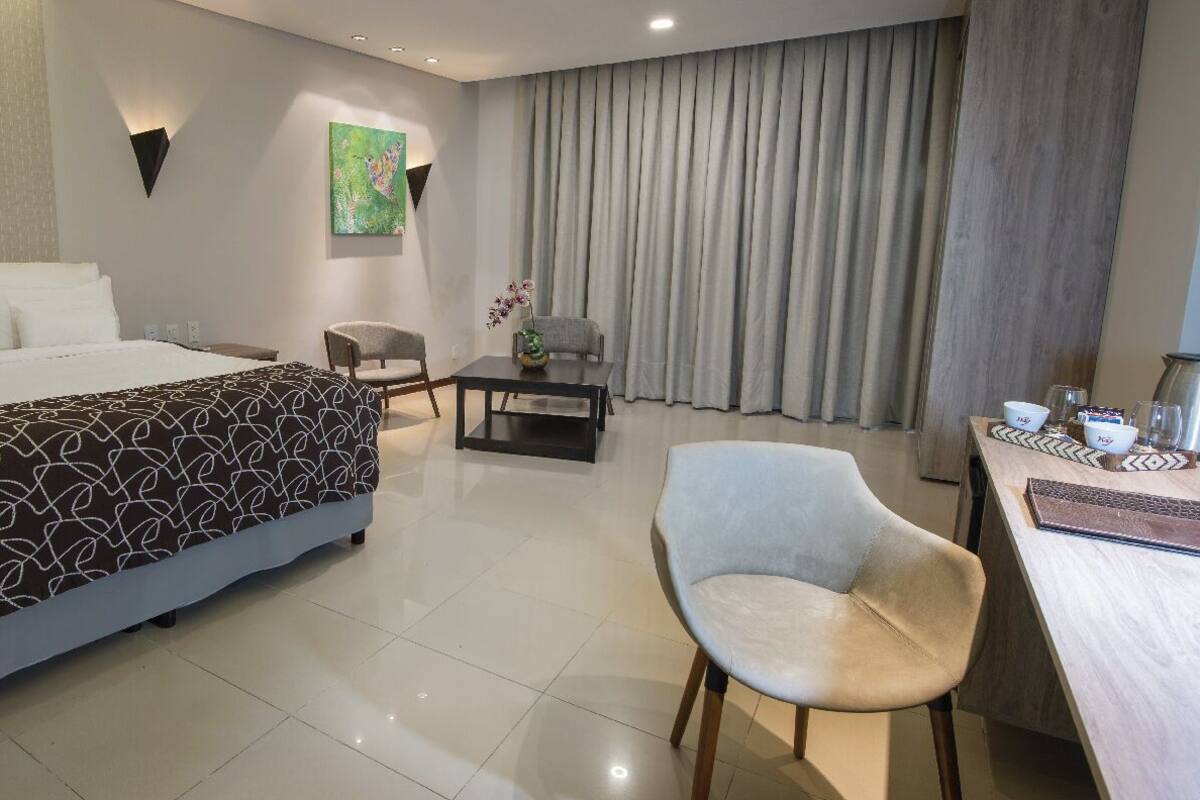 Una habitación de hotel moderna con una cama grande, un escritorio con silla, una mesita con un florero y un ventanal con cortinas.