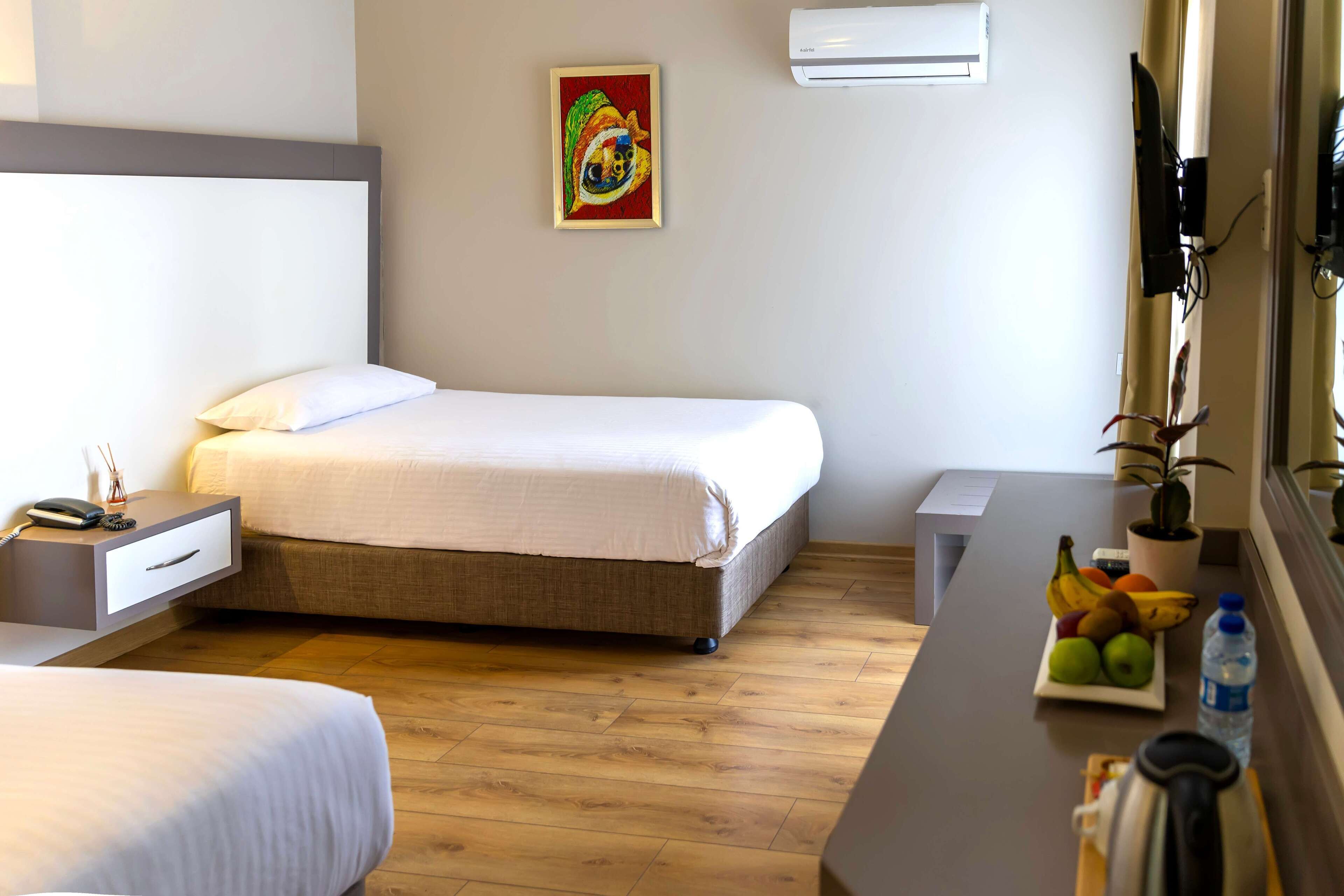 A hotel room with a bed, bedside table, a small desk, a potted plant, and a fruit bowl.