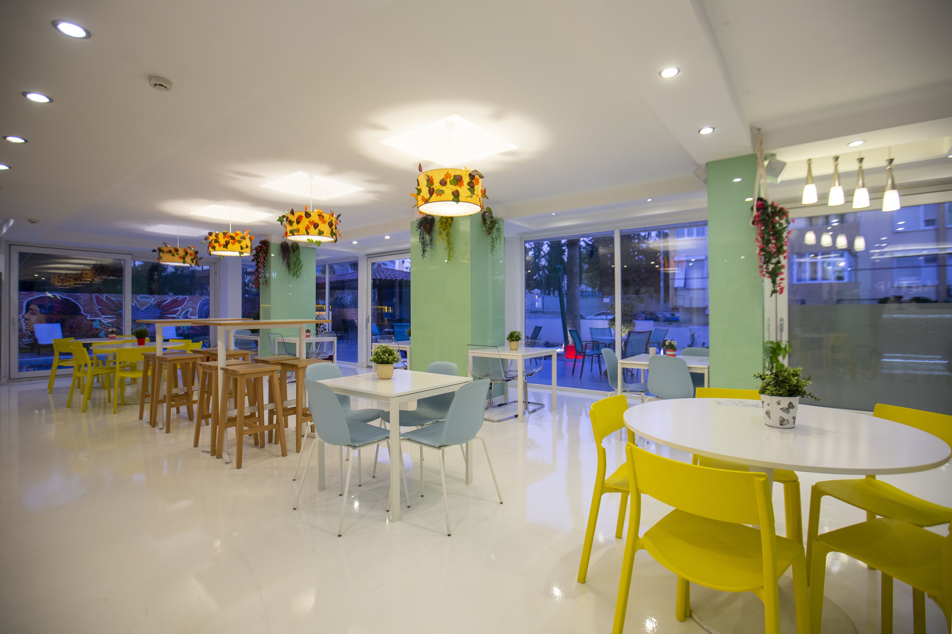 Un espace de restauration moderne avec des tables rondes, des chaises jaunes et bleues, et des piliers verts ornés de plantes suspendues.