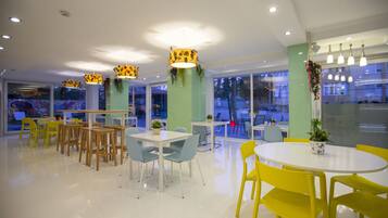 Un espace de restauration moderne avec des tables rondes, des chaises jaunes et bleues, et des piliers verts ornés de plantes suspendues.