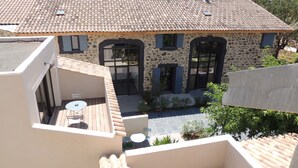 A stone building with a tiled roof, a balcony with a table and chairs, and a small garden area.