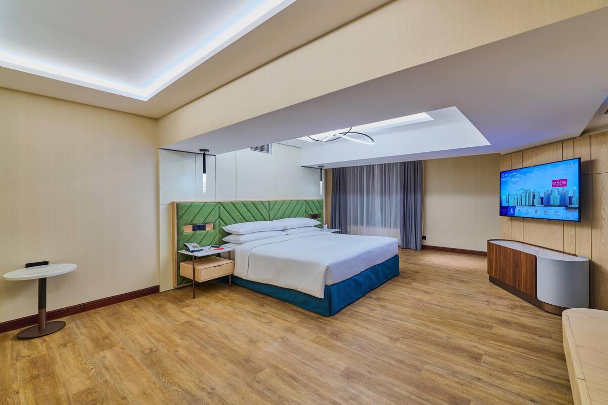 A modern hotel room with a bed, bedside table, lamp, TV, and a view of the cityscape.