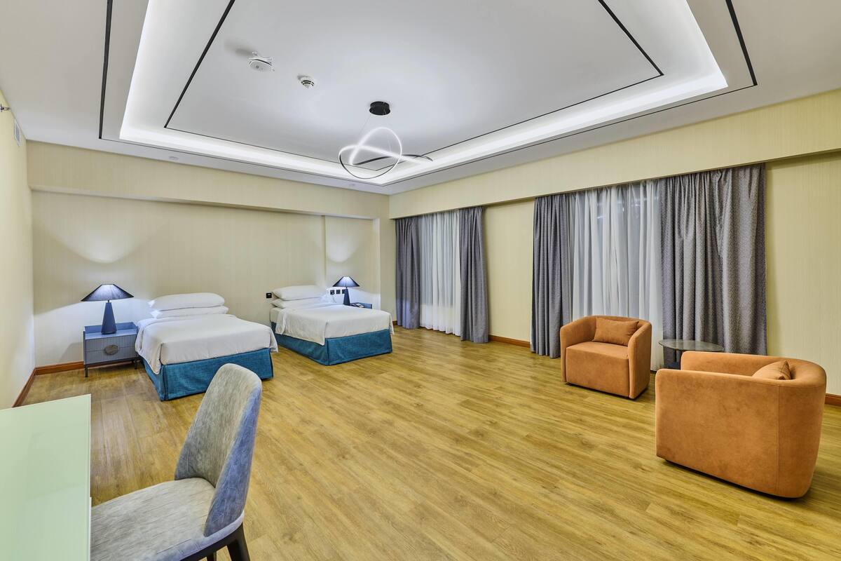 A hotel room with a wooden floor, two beds, a sofa, a chair, a desk, and a large window with curtains.