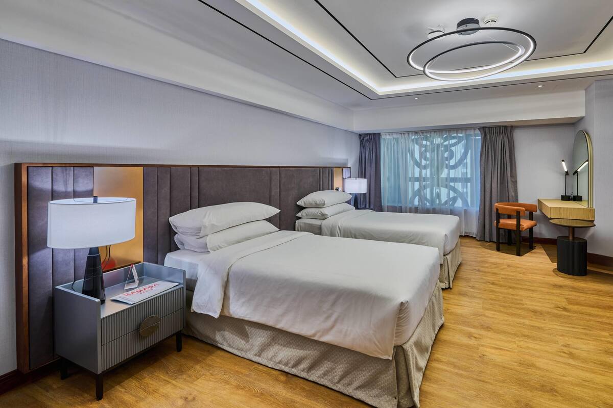 A hotel room with two beds, a nightstand, a lamp, a desk with a mirror, and a window with curtains.