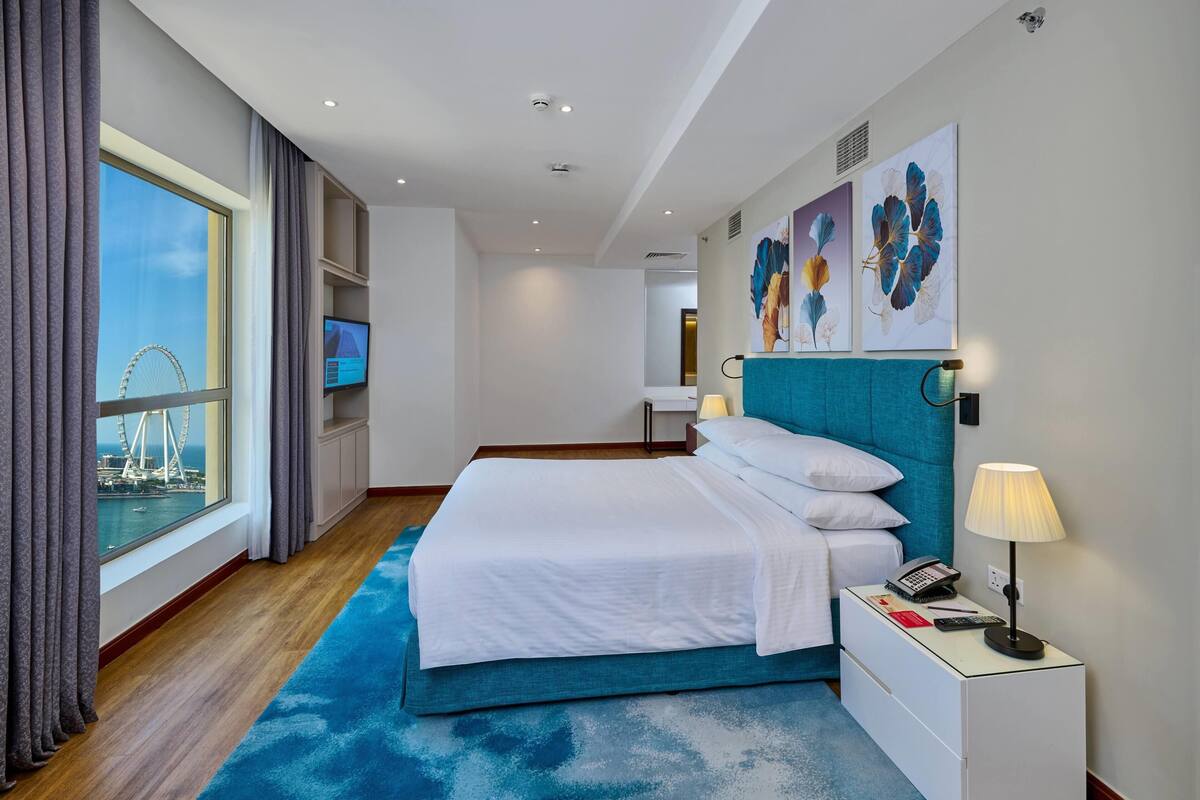 A hotel room with a large bed, a nightstand with a lamp, a TV, and artwork on the wall.