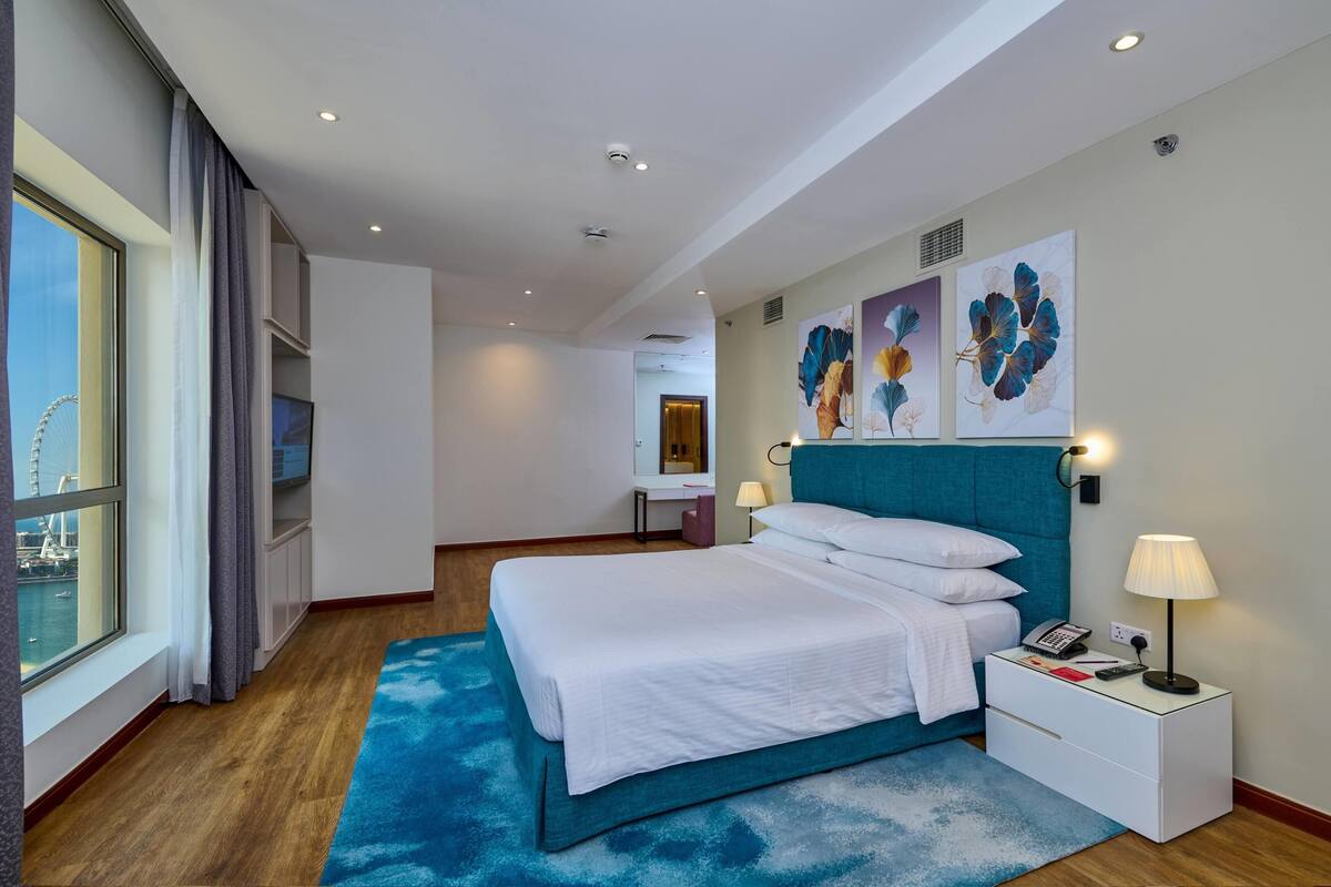 A hotel room with a bed, a nightstand, a desk, a chair, a window with curtains, and artwork on the wall.