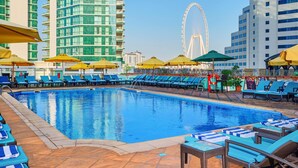 Una piscina con sillones de descanso y sombrillas, una noria al fondo y edificios altos.