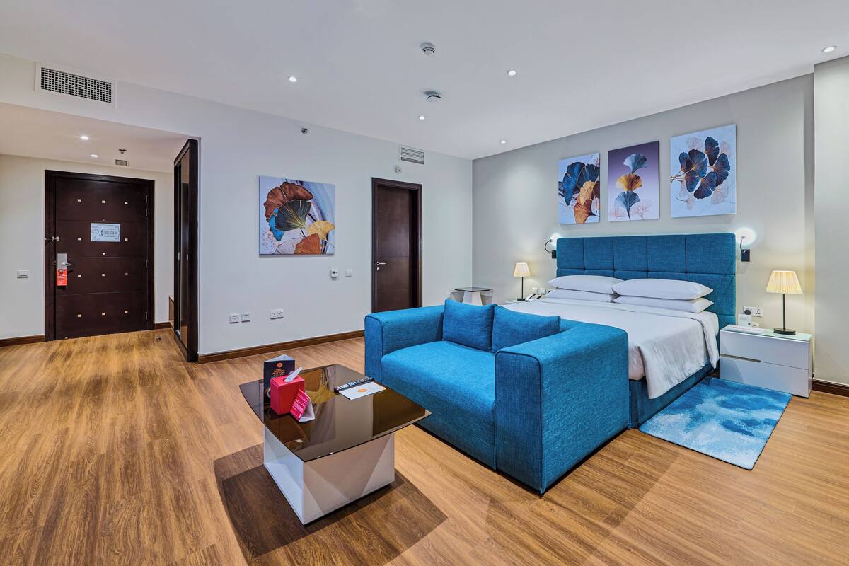A hotel room with a bed, a blue sofa, a small table, a nightstand, and artwork on the wall.