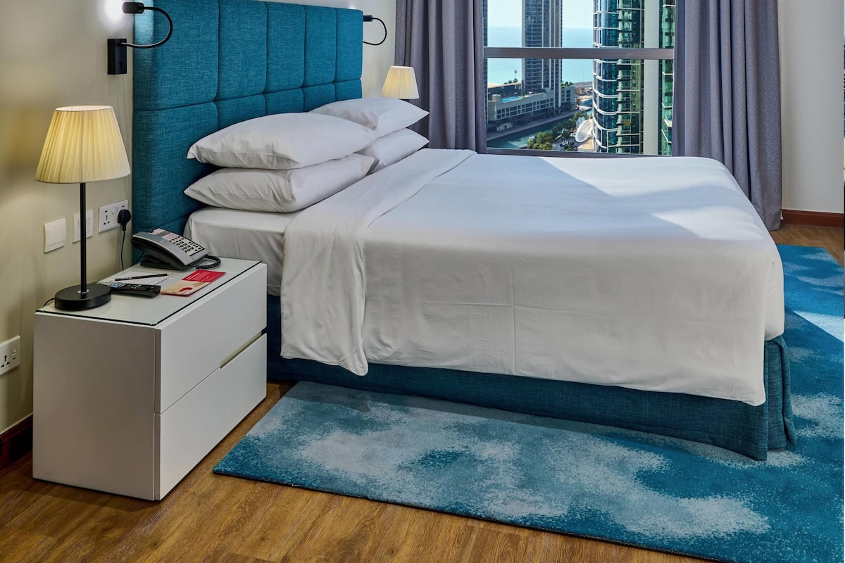 A hotel room with a bed, bedside lamps, a phone, a nightstand, and a view of the city.