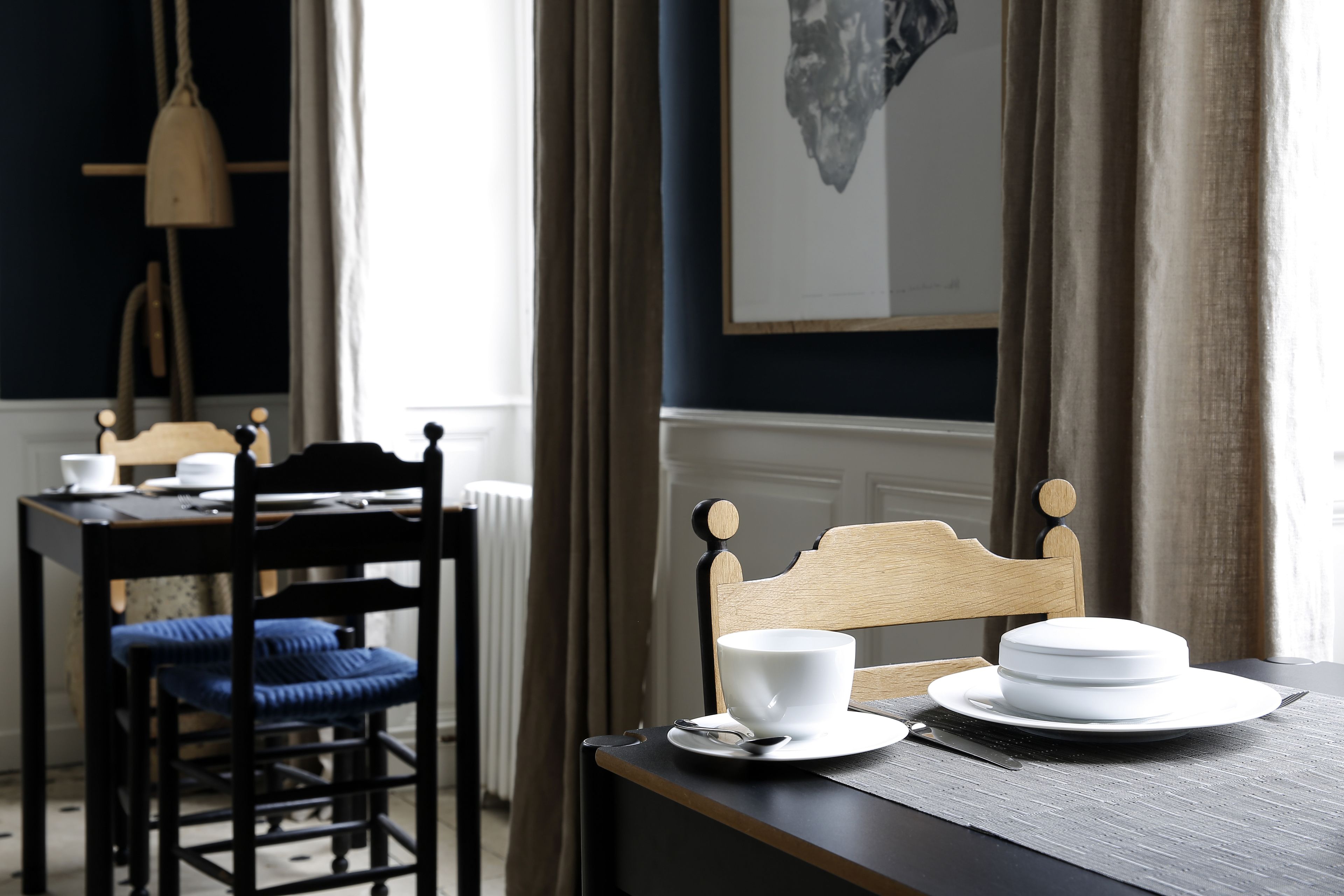 A dining area with a table set for two, featuring a white plate, a cup, and a spoon. A chair with a blue cushion is visible.