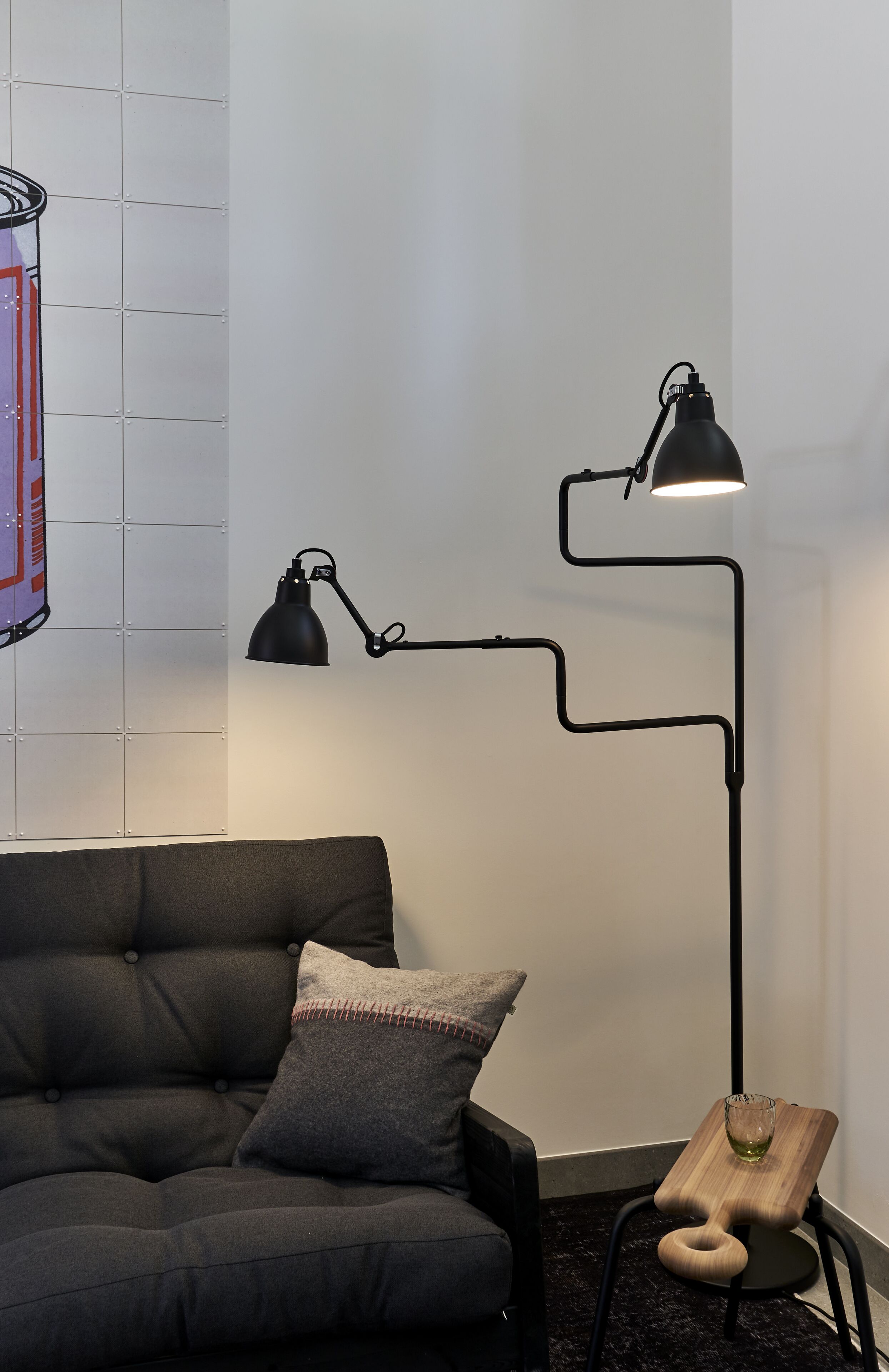 Une pièce moderne avec un lampadaire noir, un canapé foncé et une petite table d’appoint en bois sur laquelle se trouve un verre.