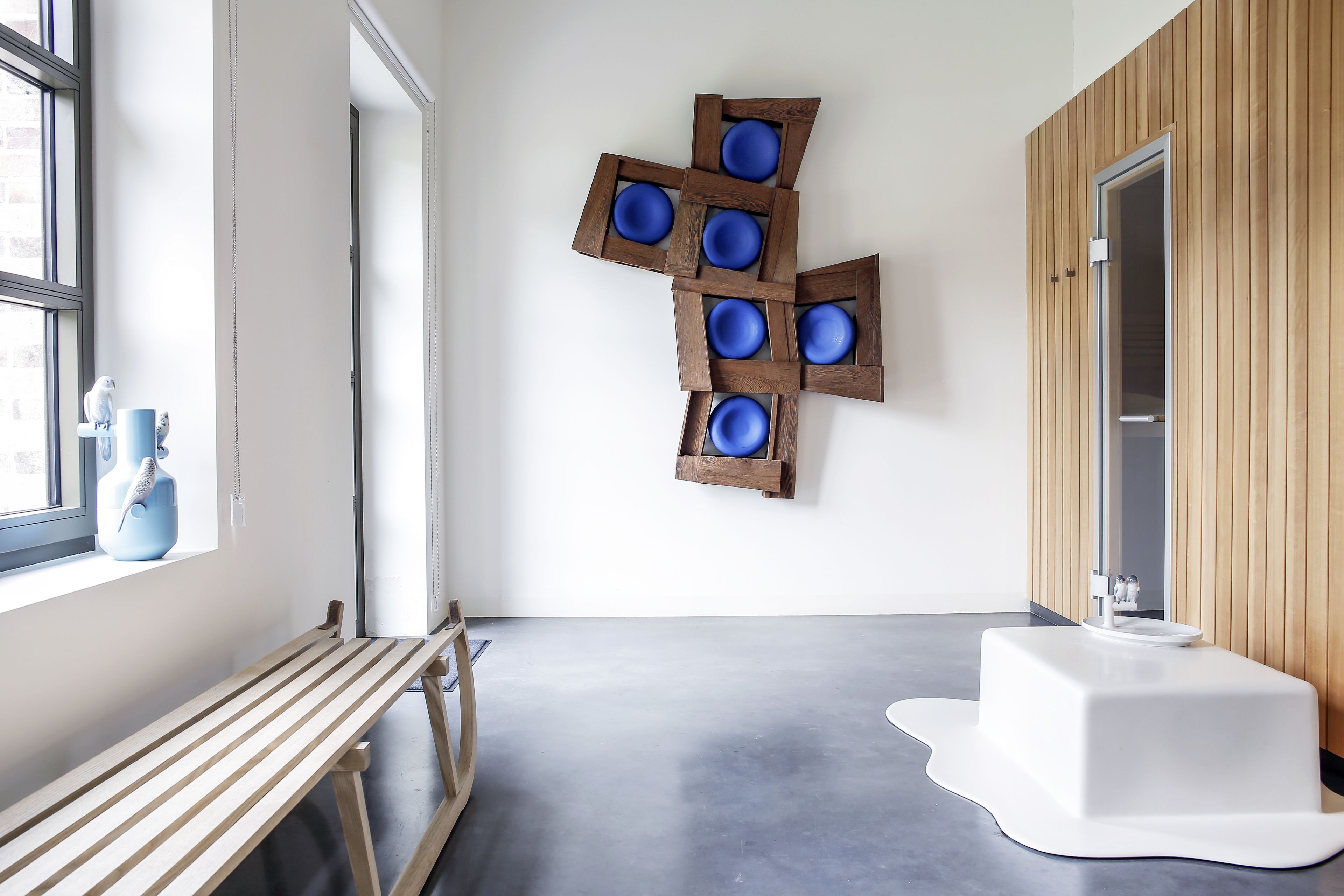 A modern room with a wooden bench, a white pedestal sink, and a wall-mounted blue bowl art piece.