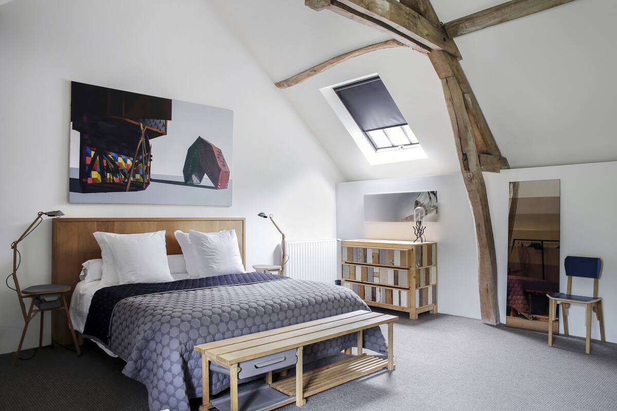 A bedroom with a sloped ceiling, a bed with a headboard, a bench, a chair, a desk, and a bookshelf.