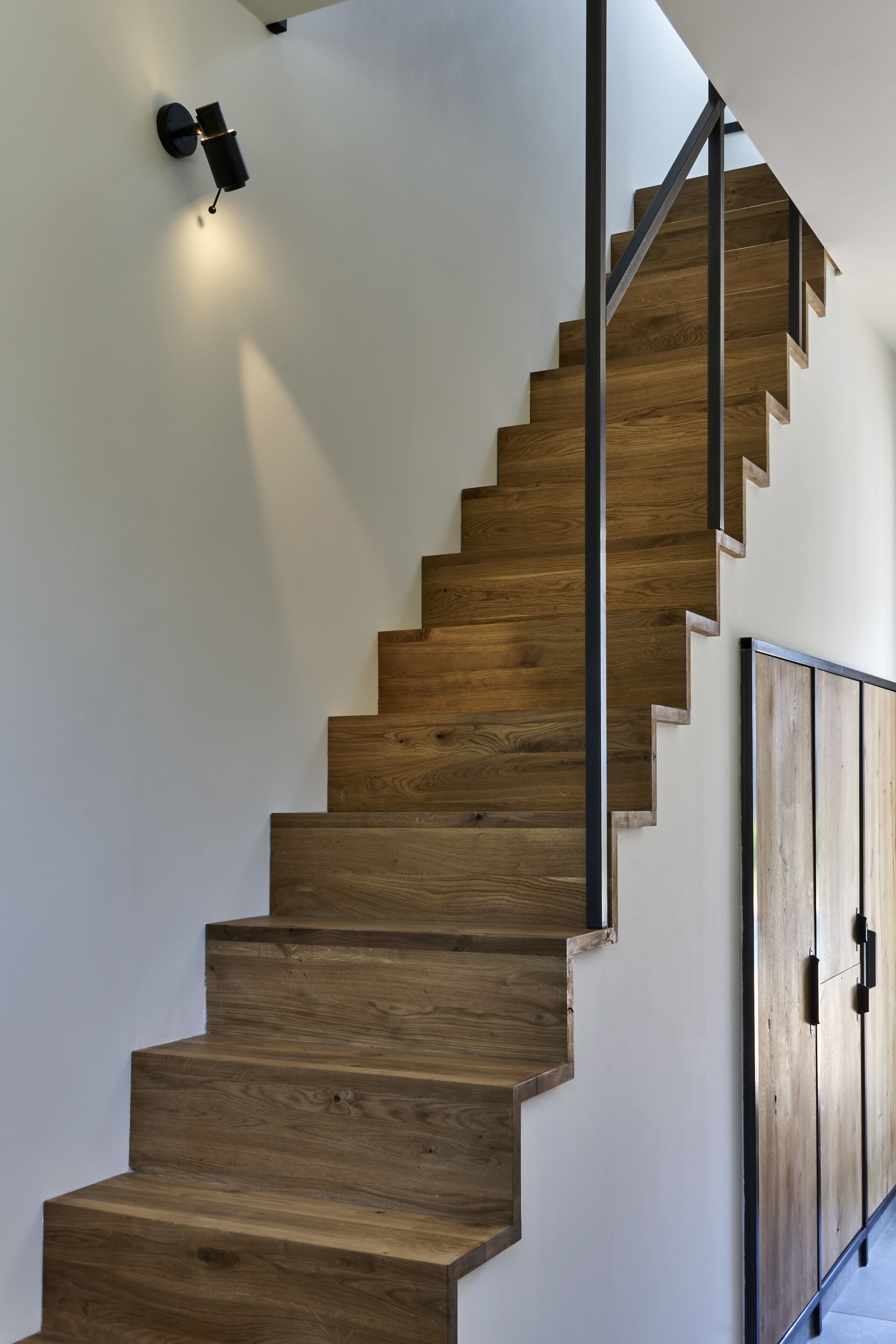 A wooden staircase with a modern design, featuring a metal handrail and a wall-mounted light fixture.