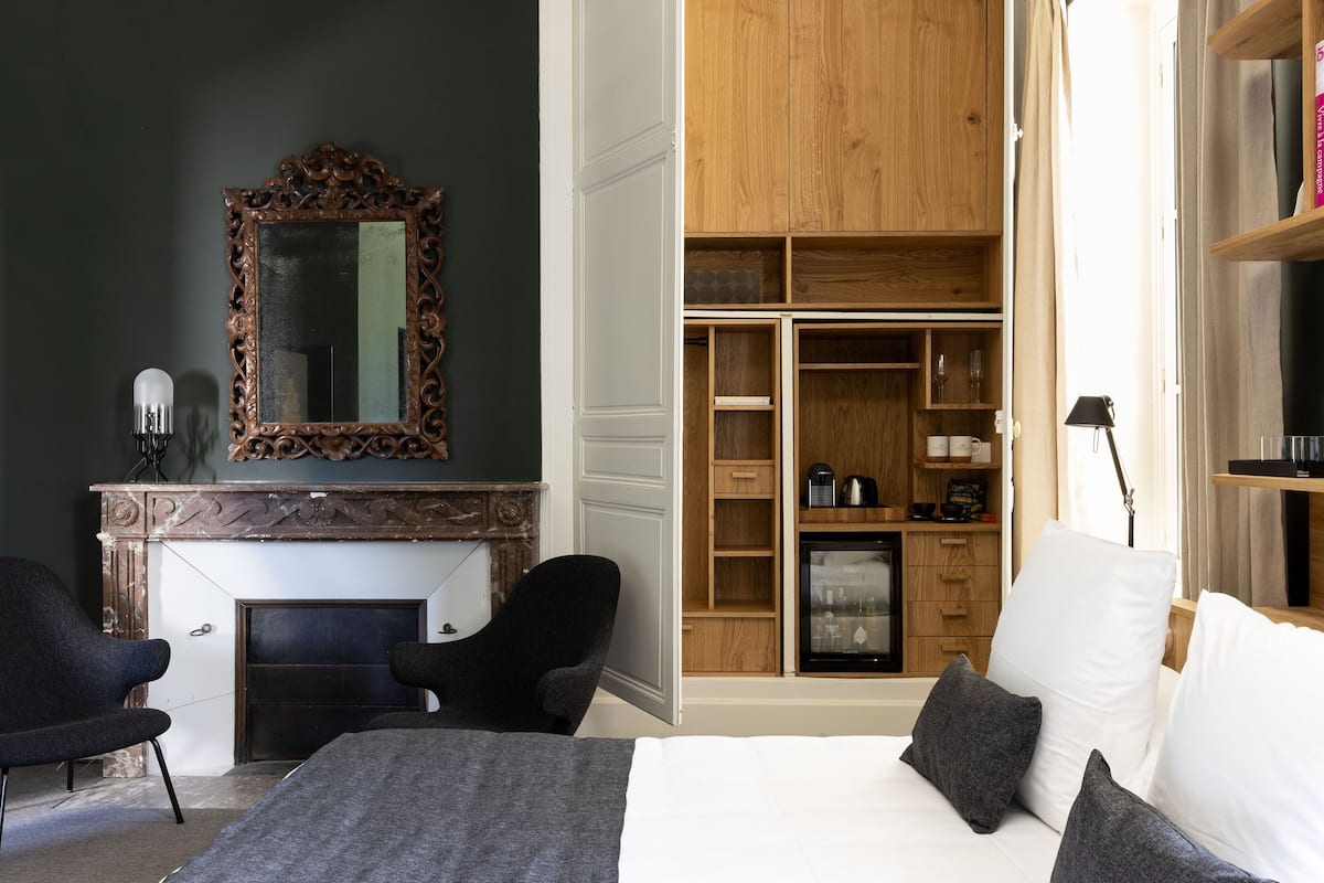 A bedroom with a bed, two chairs, a fireplace, a mirror, and a built-in wardrobe.