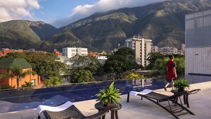 Área de piscina no terraço com espreguiçadeiras, uma mesa com planta e vista para montanhas e edifícios.
