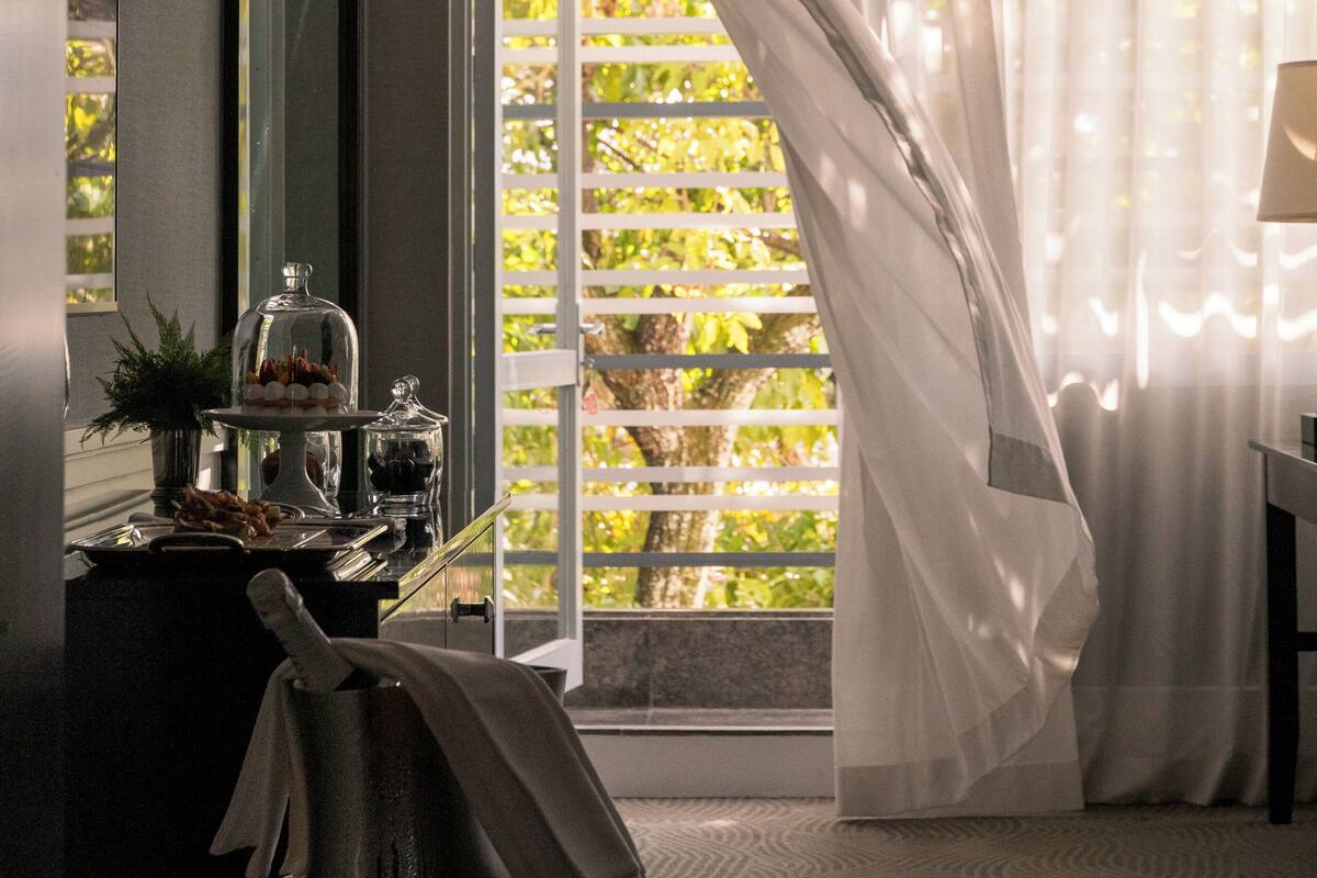Une chambre d’hôtel moderne dotée d’une grande fenêtre, d’un coin repas agrémenté d’un lustre, et offrant une vue sur la verdure environnante.
