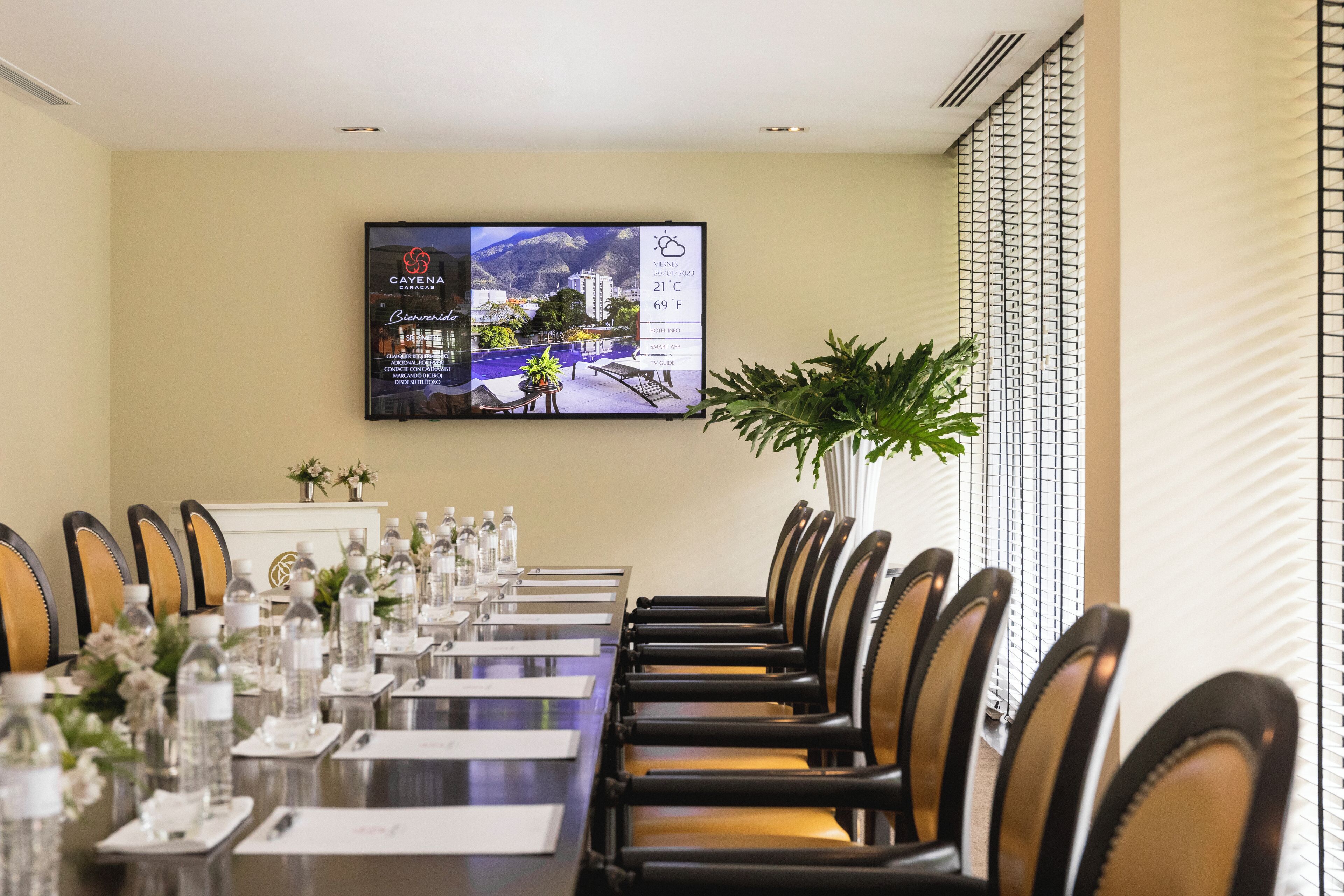 Un salón de conferencias con una larga mesa, sillas, botellas de agua y una televisión mostrando información del hotel.