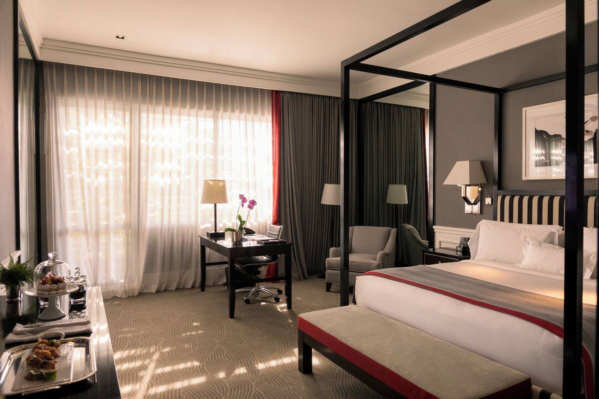 Une chambre d’hôtel avec un lit à baldaquin, un bureau avec une lampe, une chaise et une table à manger sur laquelle se trouvent un gâteau et des fruits.
