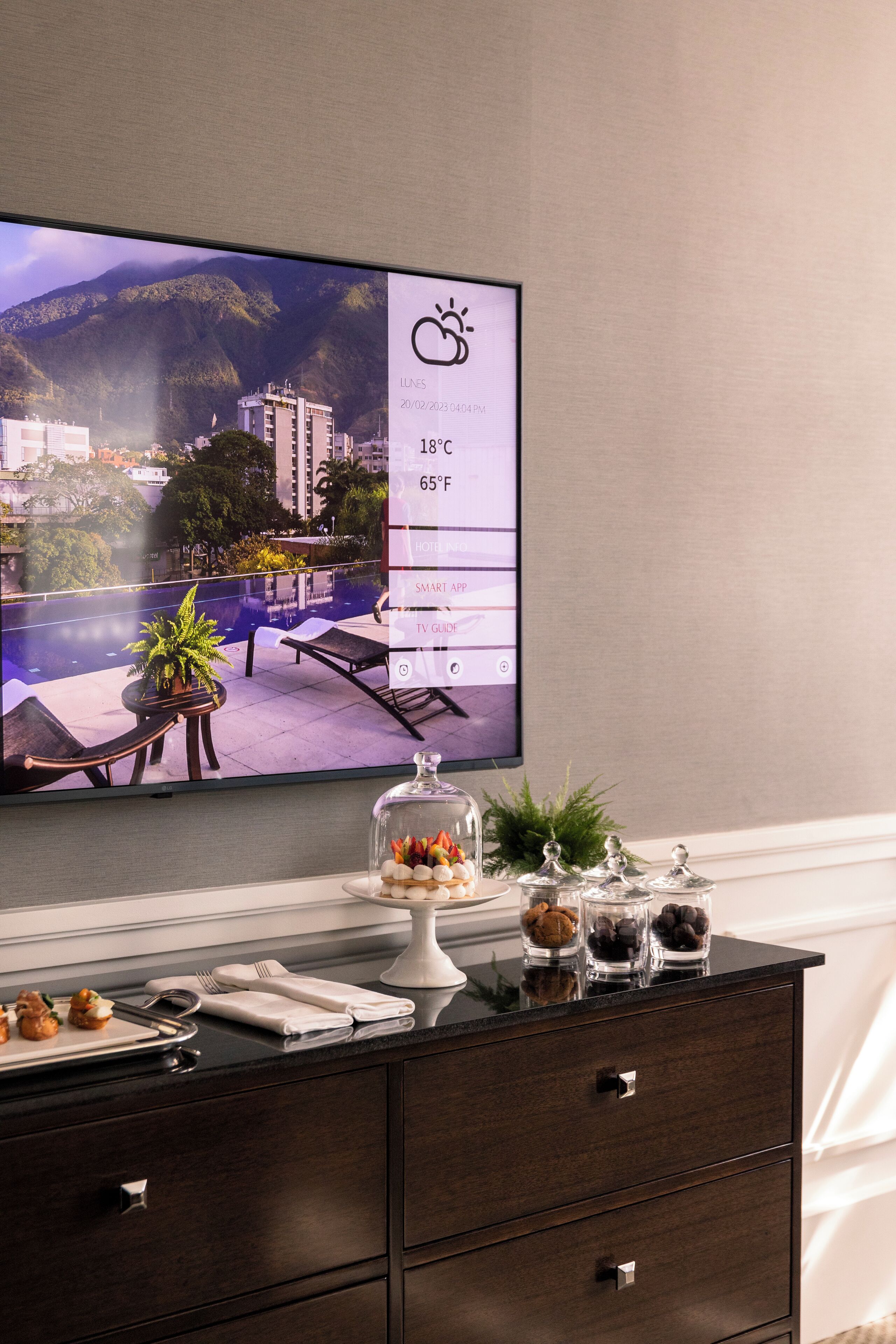 A modern hotel room with a large TV displaying a scenic mountain view, a dark wooden dresser with a glass display case, and neatly arranged amenities.