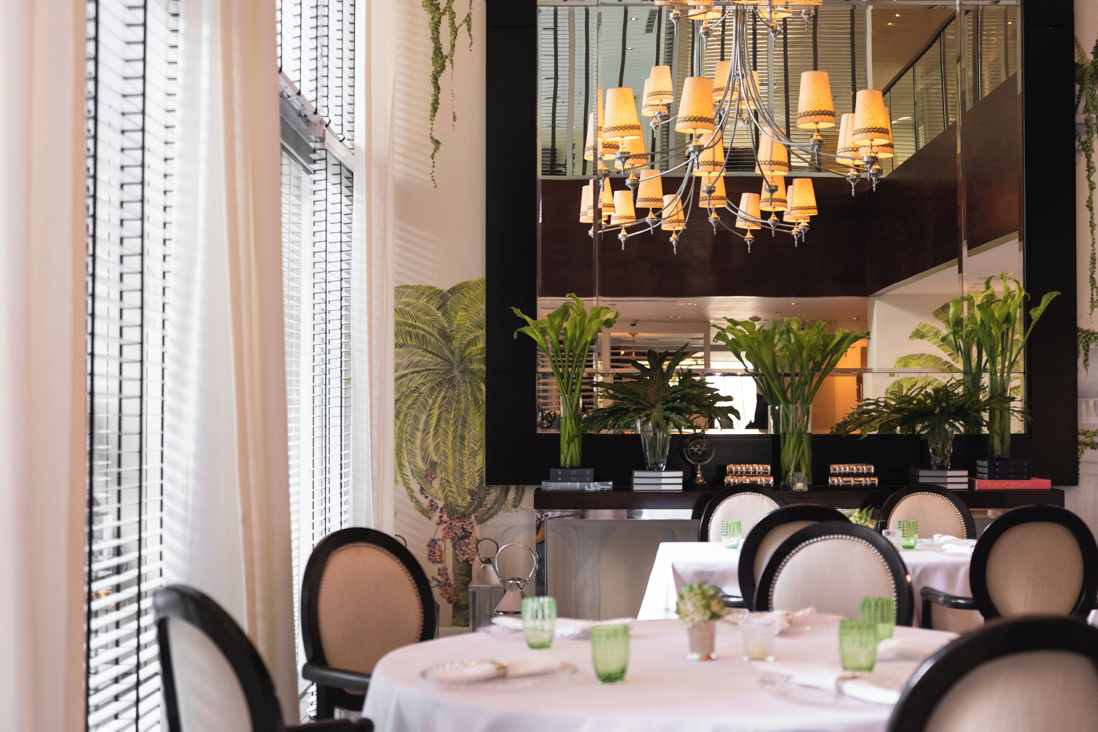 A dining area with a chandelier, tables set with green glassware, and potted plants.