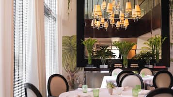 A dining area with a chandelier, tables set with green glassware, and potted plants.