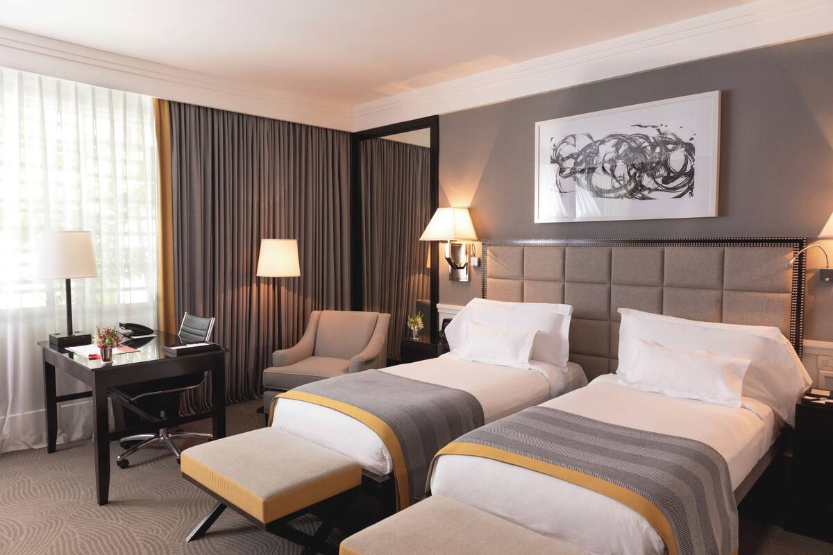 Une chambre d’hôtel avec deux lits, un bureau, une chaise, une lampe et un tableau au mur.