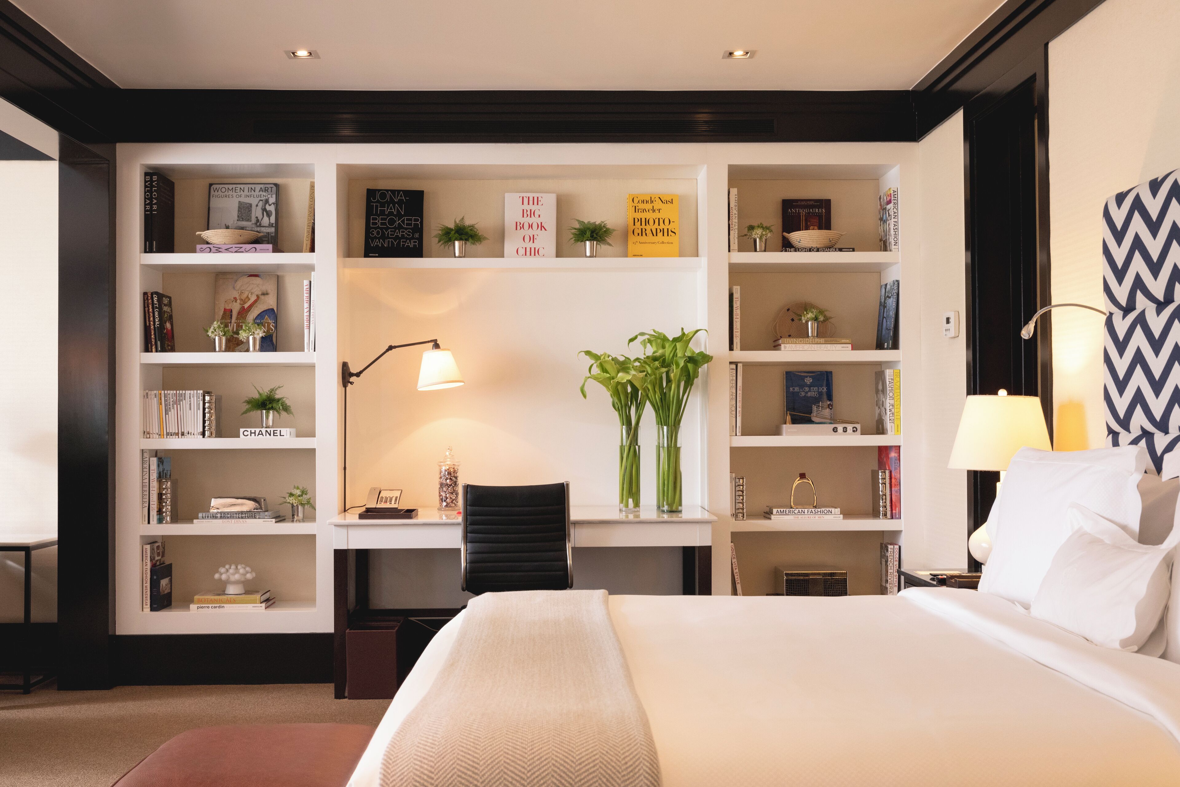 A hotel room with a large bed, a desk with a chair, and a bookshelf filled with books and decorative items.