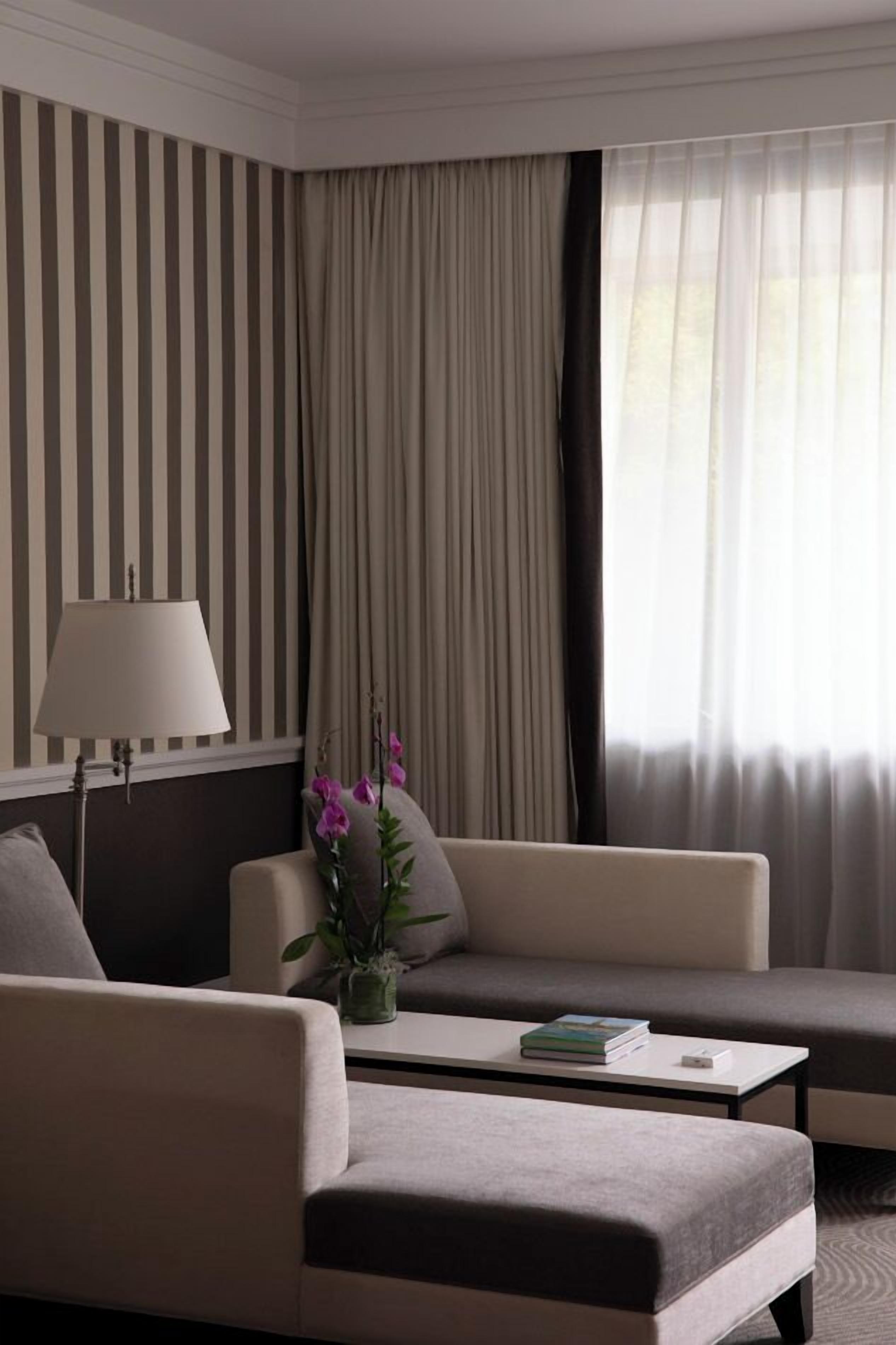 A hotel room with striped wallpaper, a sofa, a coffee table, a lamp, and a window with curtains.