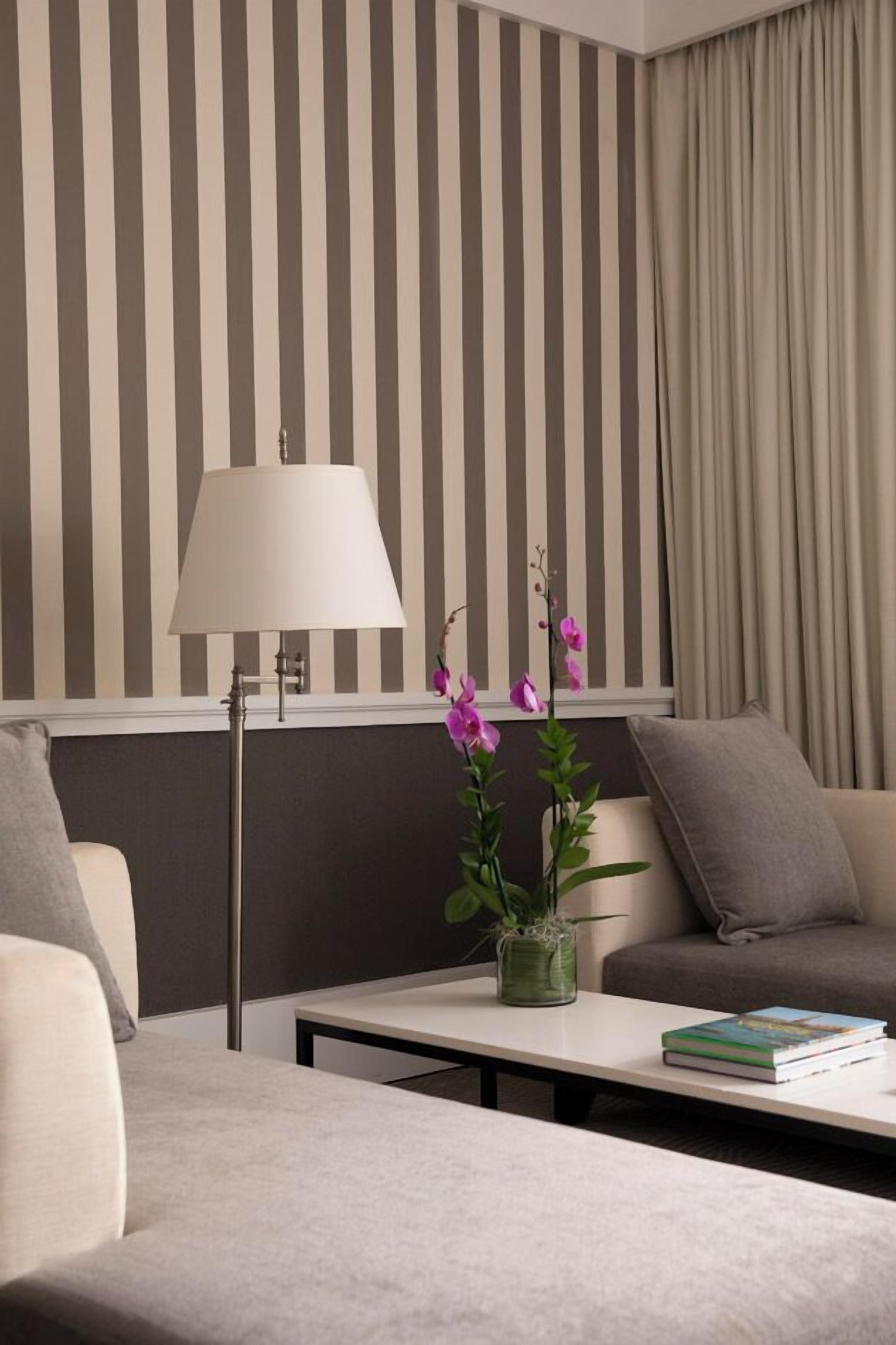 A modern hotel room with striped wallpaper, a lamp, a small table with a book, and a plant.