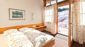 A bedroom with a wooden headboard, a bed with floral bedding, a bedside table, a framed picture on the wall, and a view of a snowy landscape through a window.