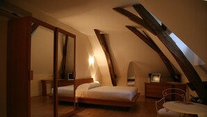 Une chambre avec un plafond en pente, des poutres en bois, un lit, un bureau, une chaise et une petite table avec une lampe.