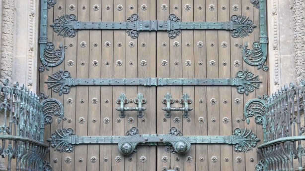 Une grande porte en bois richement décorée, ornée de motifs métalliques complexes et surmontée d’un bas-relief en pierre.