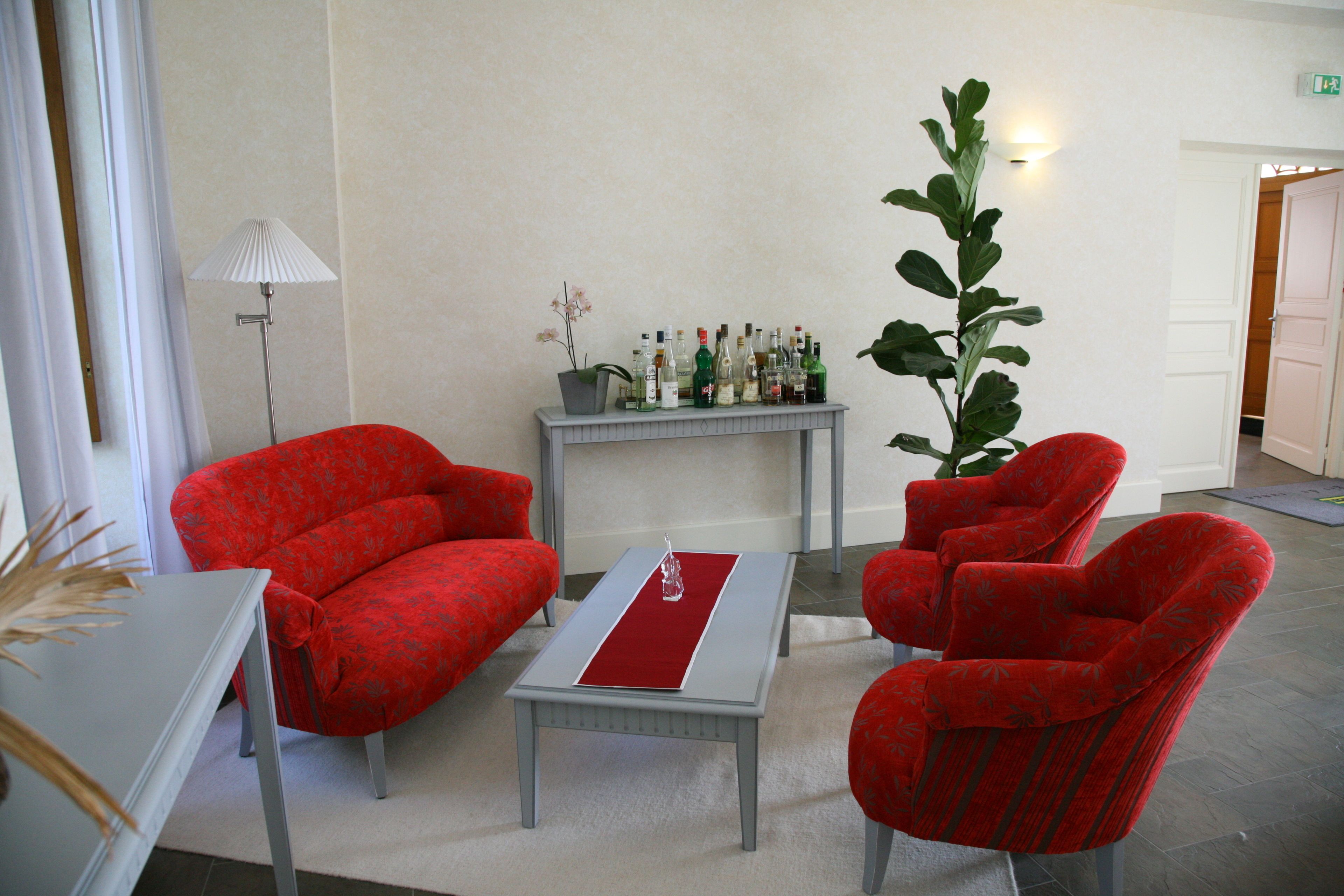 A hotel room with red upholstered chairs, a small table with a red runner, a potted plant, and a shelf with various bottles.