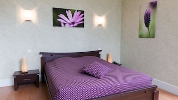 Una cama bien hecha con una colcha morada estampada y almohadas, flanqueada por dos mesitas de noche con lámparas, frente a una pared con dos cuadros de flores.