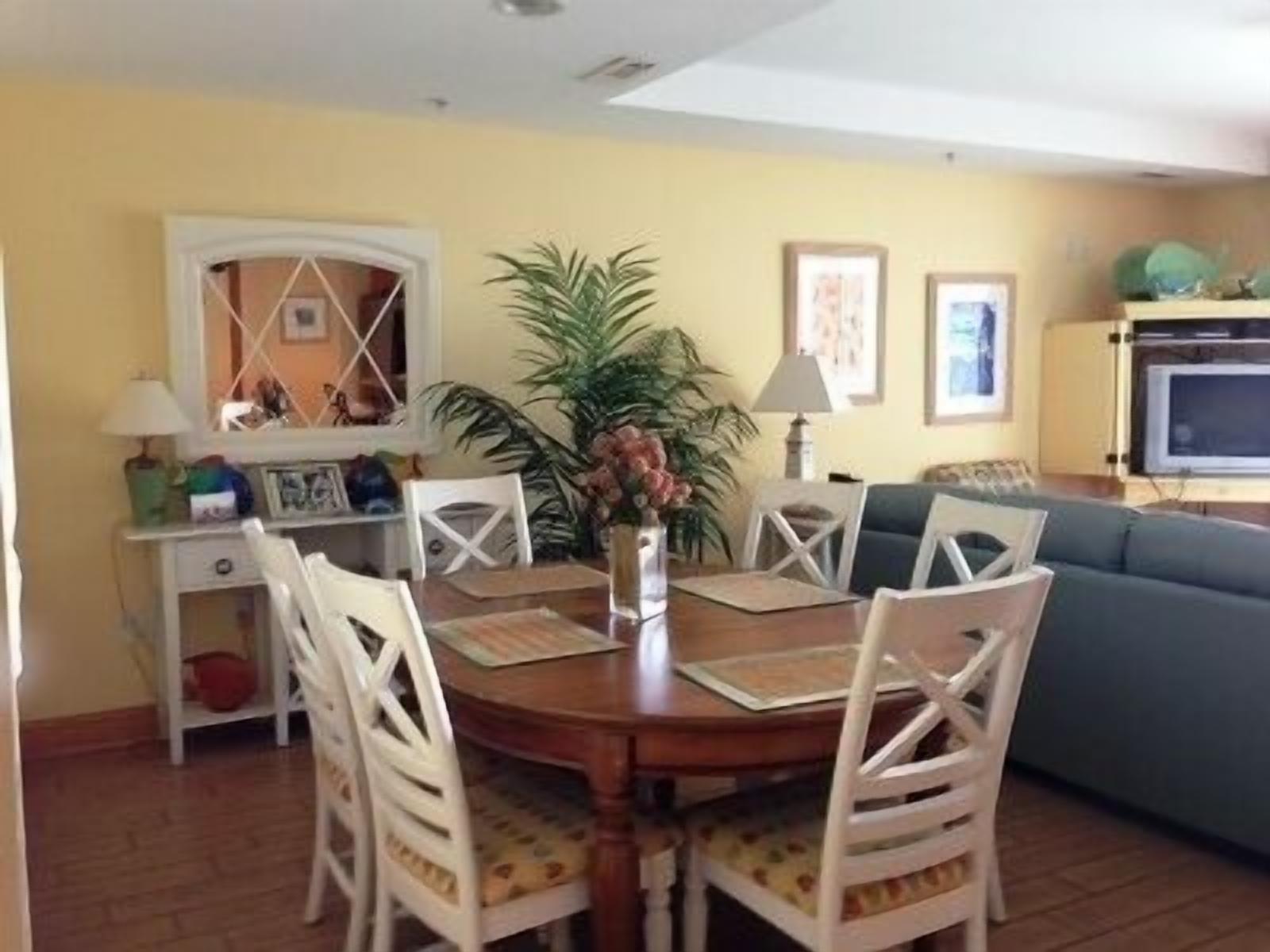 A dining area with a wooden table, white chairs, a vase with flowers, and a green sofa in the background.