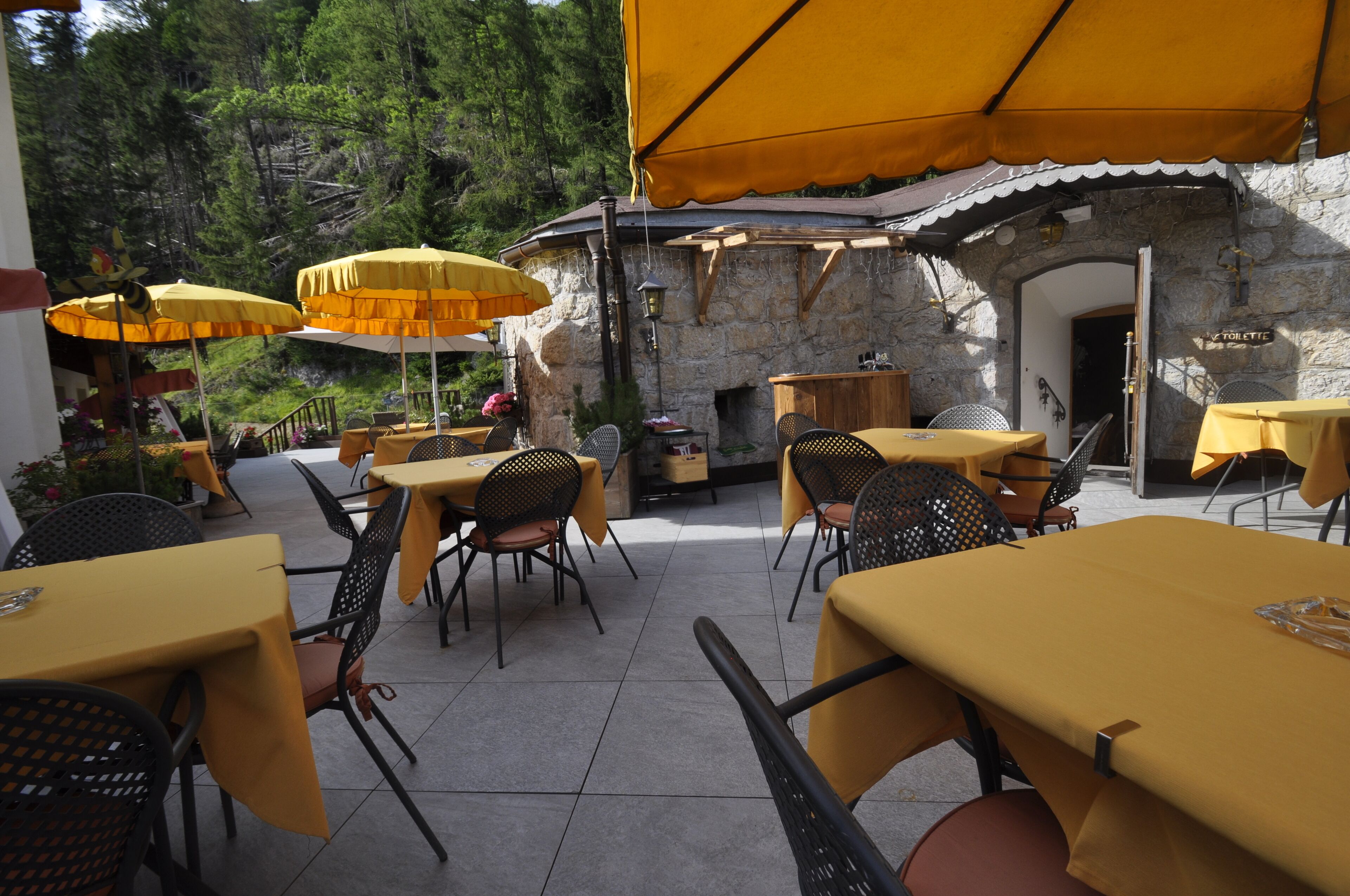 An outdoor café with yellow tables and chairs, stone walls, and umbrellas providing shade.