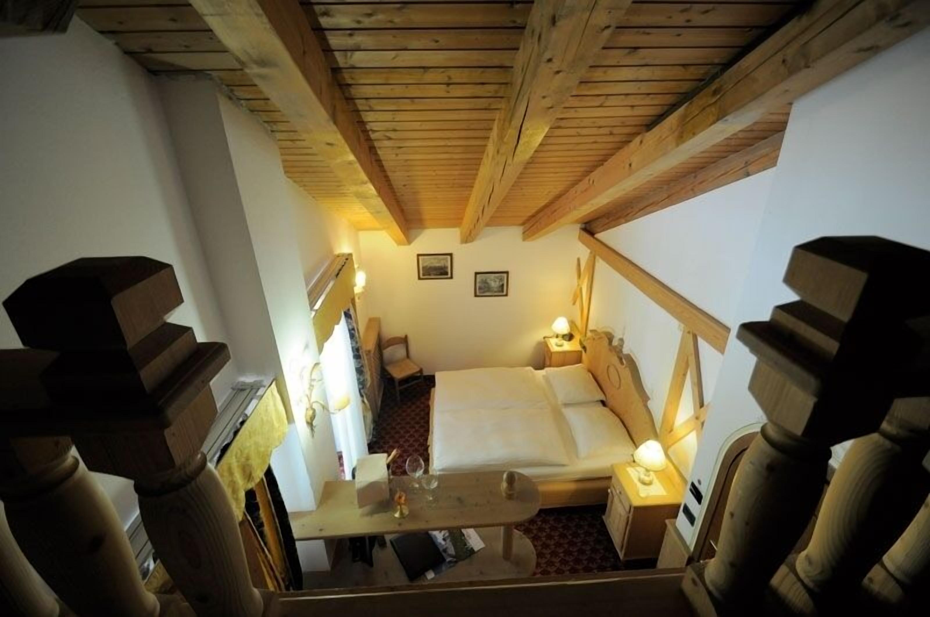 A small, cozy room with a wooden ceiling, a bed, a nightstand with a lamp, and a small table with a glass.