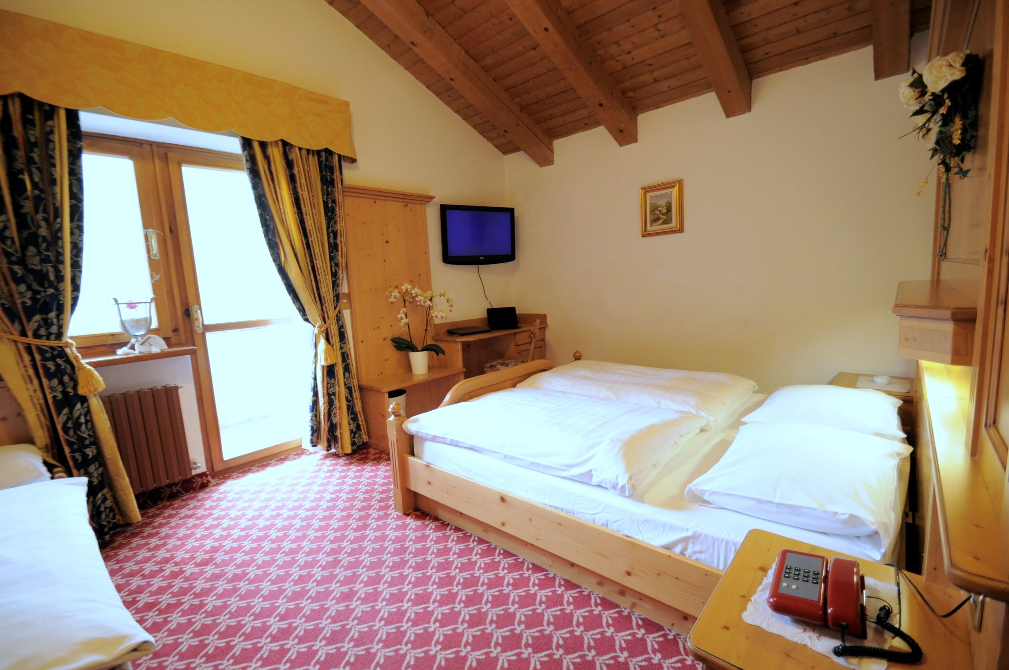 Habitación de hotel con una cama de madera, un televisor montado en la pared, un teléfono en una mesita de noche y una ventana con cortinas.