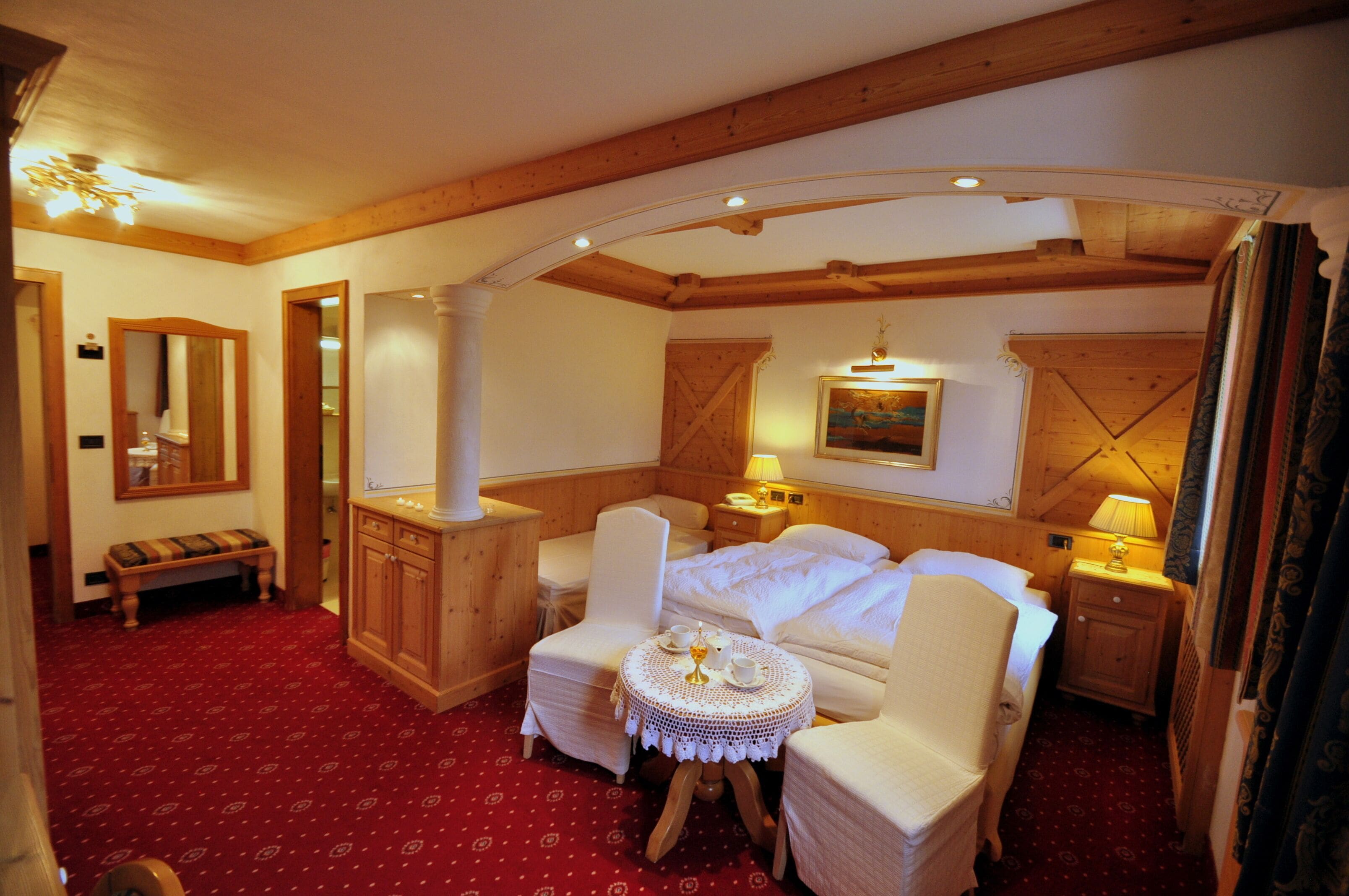 A cozy hotel room with a wooden ceiling, a large bed, two bedside tables with lamps, a small round table with a decorative centerpiece, and a bench in the corner.