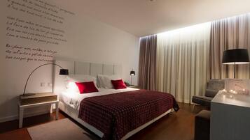 Una habitación de hotel moderna con una cama bien hecha, una mesita de noche con lámpara, un escritorio con lámpara y un amplio ventanal con cortinas.