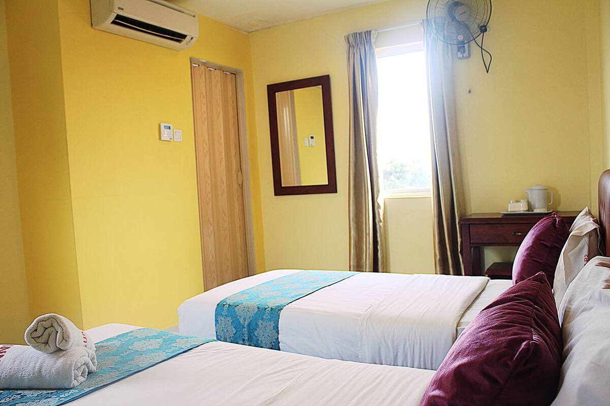 A hotel room with two beds, a window with curtains, an air conditioning unit, a mirror, and a bedside table with a cup.