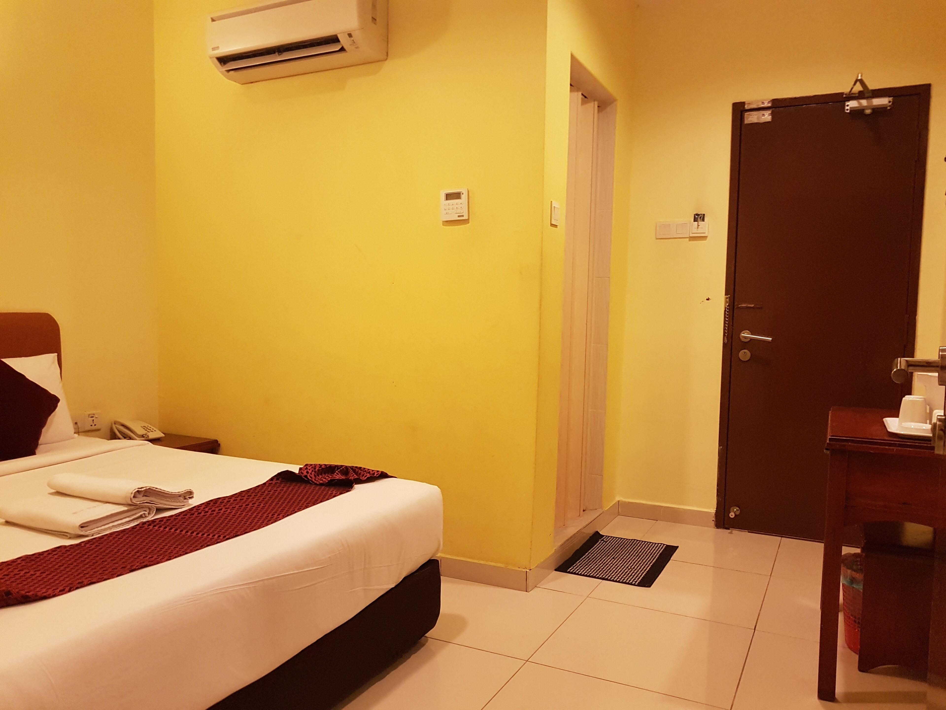 A hotel room with a bed, a bedside table with a phone, a wall-mounted air conditioner, a door, and a small mat near the door.