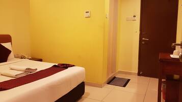 A hotel room with a bed, a bedside table with a phone, a wall-mounted air conditioner, a door, and a small mat near the door.