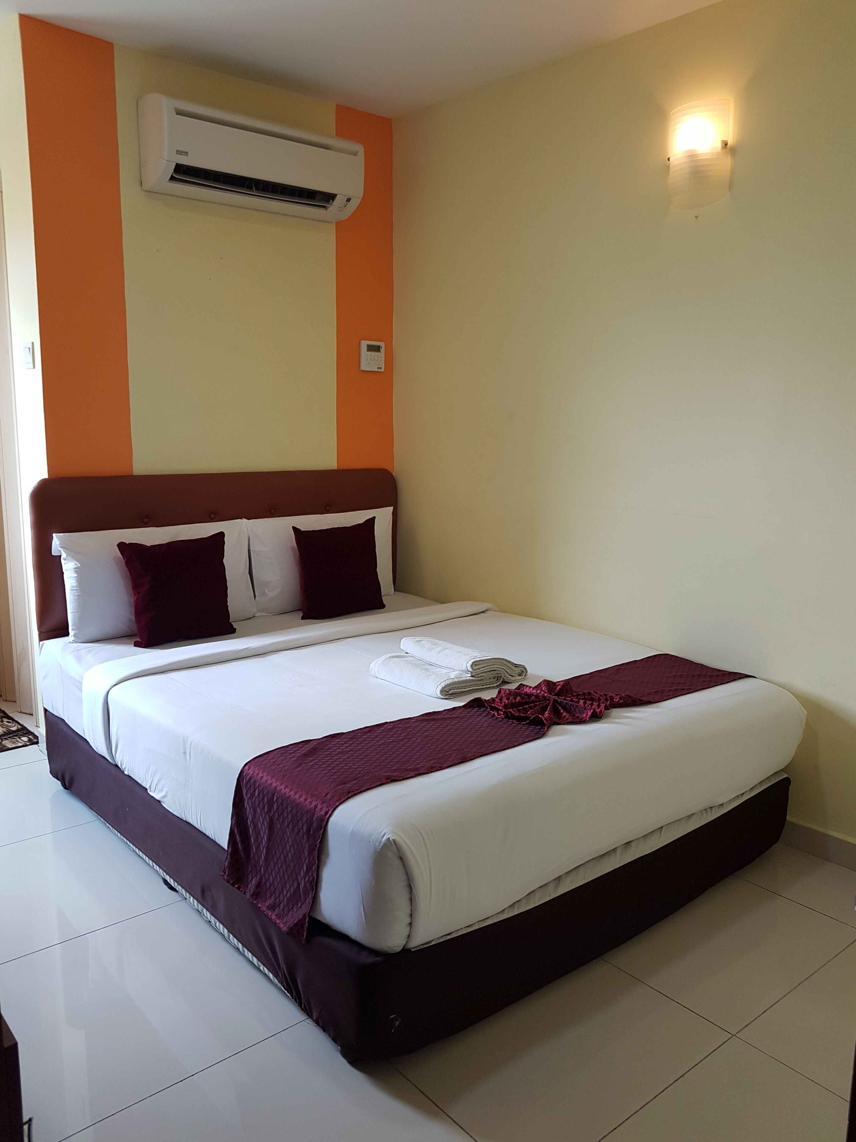 A neatly made bed with white linens and a dark brown headboard, an air conditioning unit mounted on the wall, and a wall-mounted light fixture.