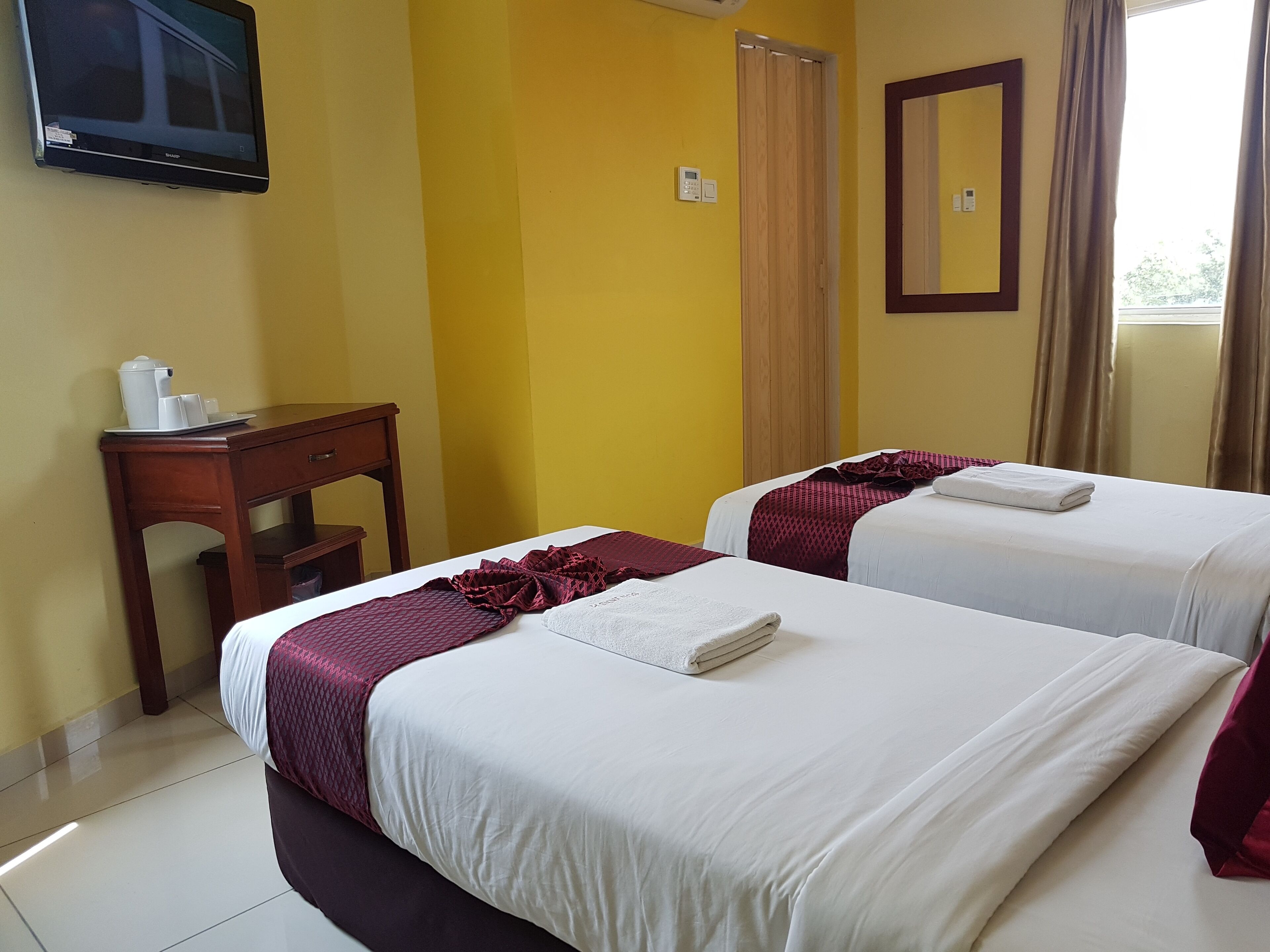 A hotel room with two beds, a wooden desk, a television mounted on the wall, and a window with curtains.