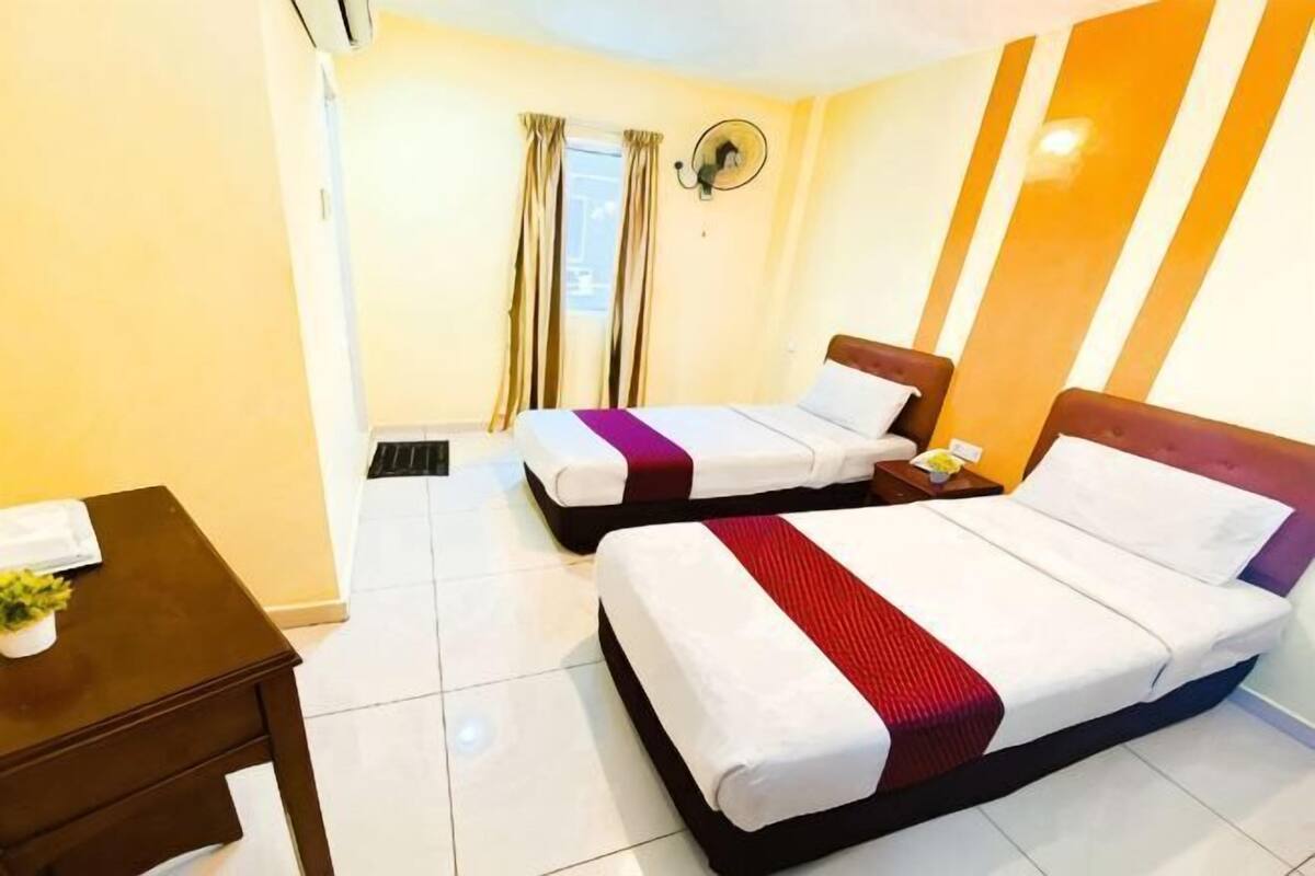 A hotel room with two beds, a small table, a wall-mounted heater, and a window with curtains.